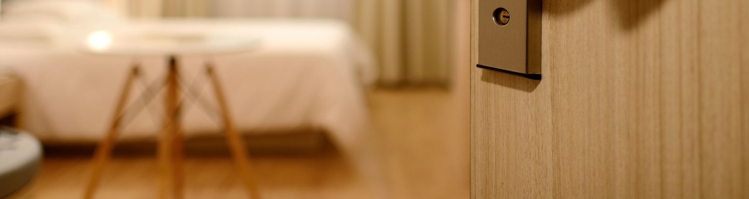 Eine Hand öffnet eine Hotelzimmertür und gibt den Blick frei auf ein wohliges Zimmer