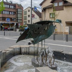 Im Vordergrund der Erzählstein aus Bronze mit mehreren kleinen Wasserfontänen und im Hintergrund die Farmstraße