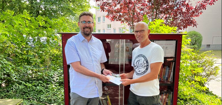 Erster Stadtrat Karsten Groß überreicht Michael Geiß einen Gutschein. Beide stehen lächelnd vor einem Bücherschrank